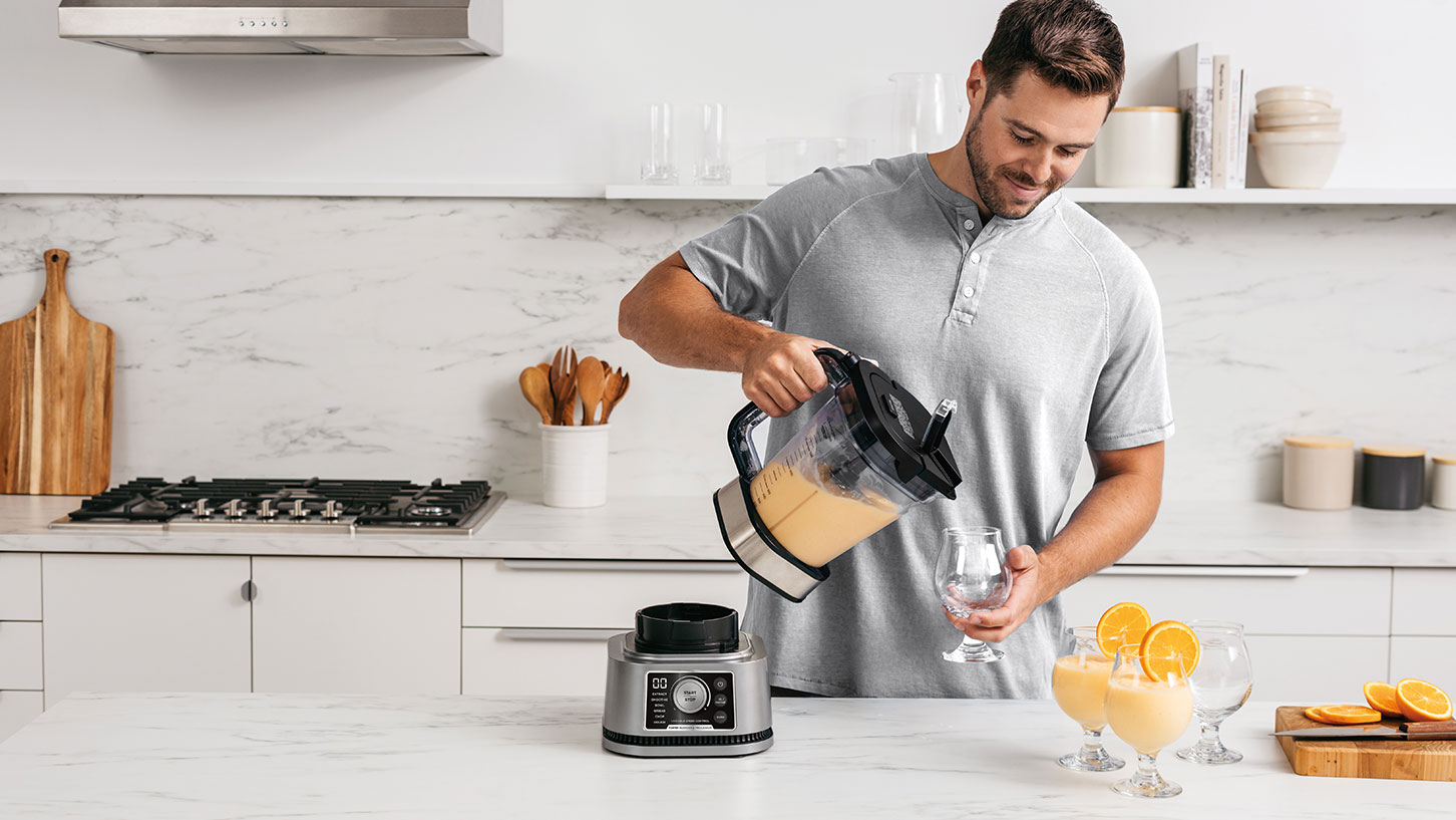 Man in a kitchen pouring out an orange smoothie into glasses from a blender